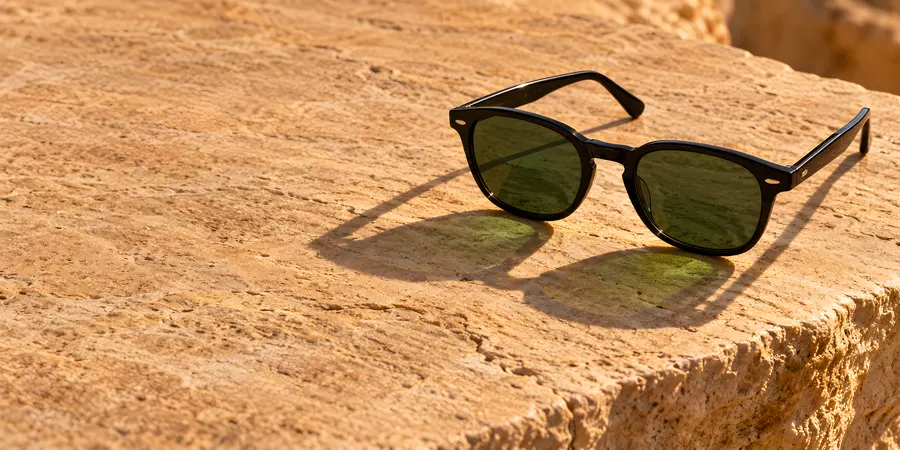 Schwarze Sonnenbrille mit grünen Gläsern auf warmem Sandstein in tiefer Abendsonne, ein grünlicher Lichtfleck fällt durch die Gläser
